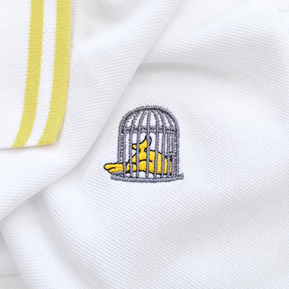 Close-up of Sauce & Brown Capri white polo showing yellow cuff trim and embroidered birdcage logo.