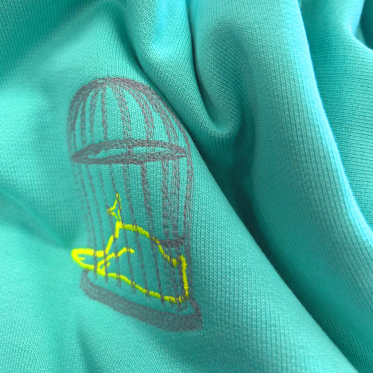Close-up of Sauce & Brown Pool sweatshirt showing embroidered birdcage chest logo detail