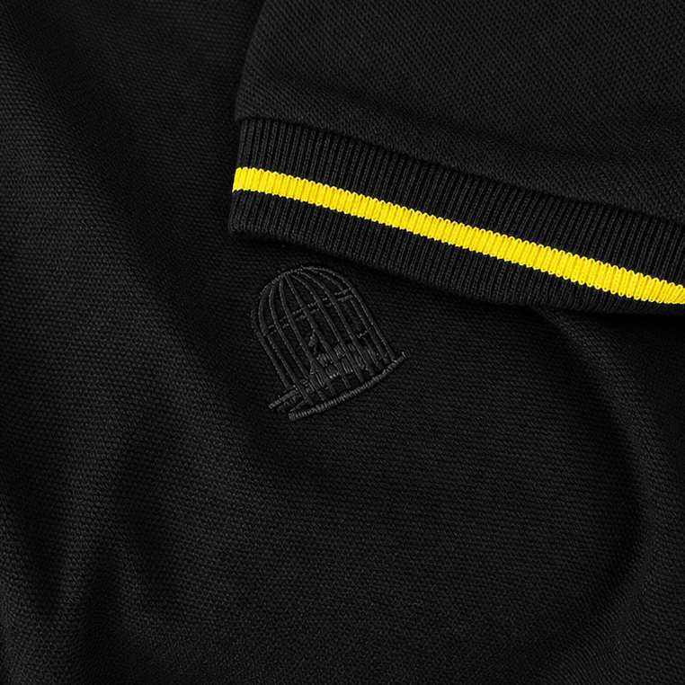 Close-up of Sauce & Brown Offset black polo showing the yellow cuff stripe and tonal embroidered birdcage logo.